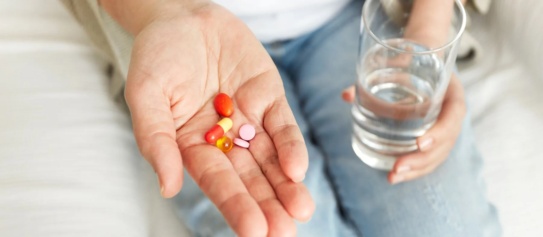 An open palm holding several tablets and capsules, with a glass of water in the other hand