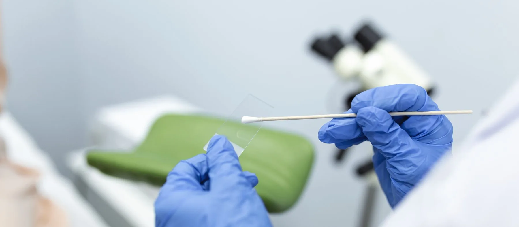 A clinician’s gloved hands holding a swab and slide in an examination room