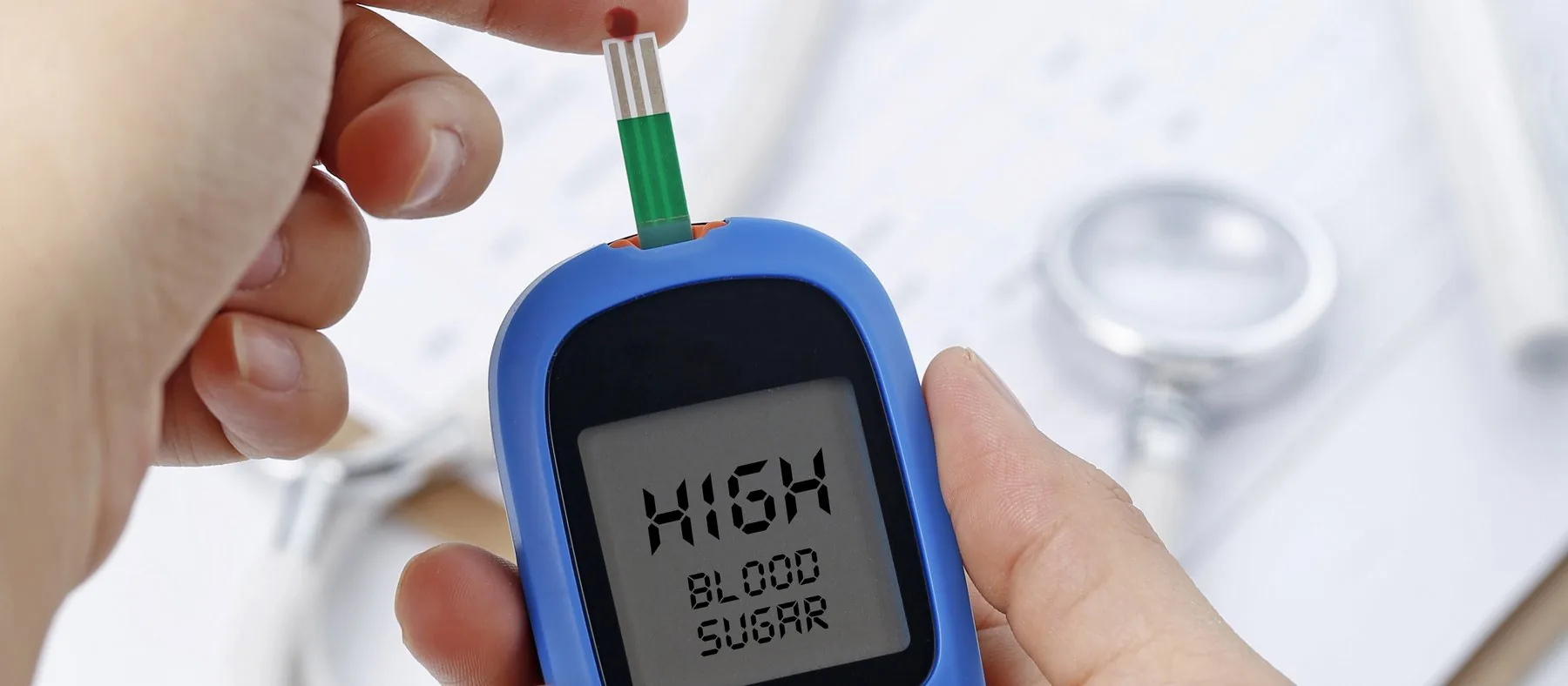 A hand holding a blood glucose meter, with a stethoscope and chart behind it