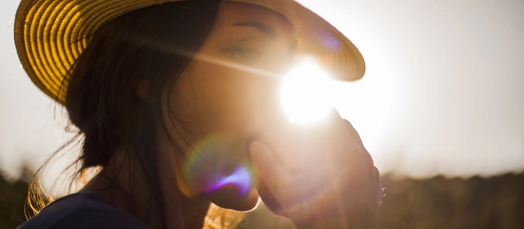 A woman in a wide-brimmed straw hat shading her eyes from low, bright sun
