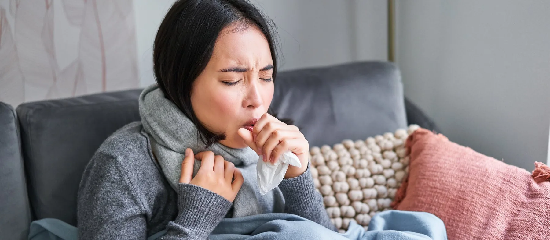 A woman wrapped in a blanket and scarf coughing into a tissue at home